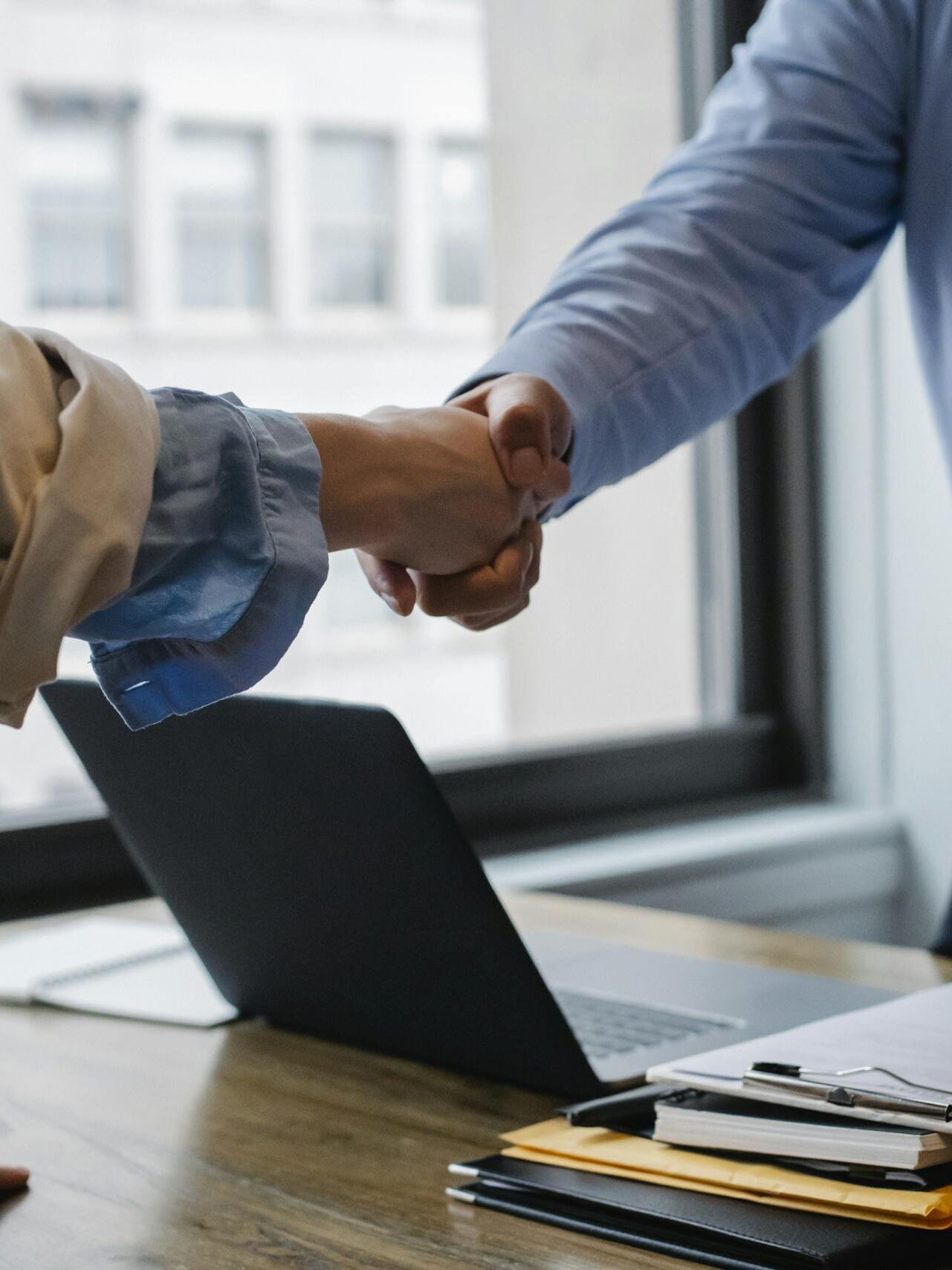 Crop unrecognizable coworkers in formal wear standing at table with laptop and documents while greeting each other before meeting