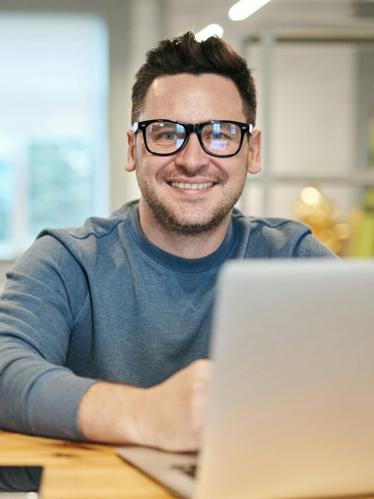 Happy man wearing glasses working remotely on laptop in modern office environment.